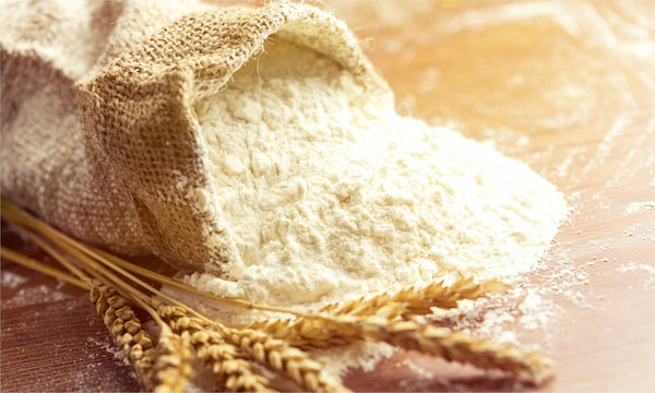 A brown bag of flour spills out artistically on a table. There are stalks of wheat next to it.