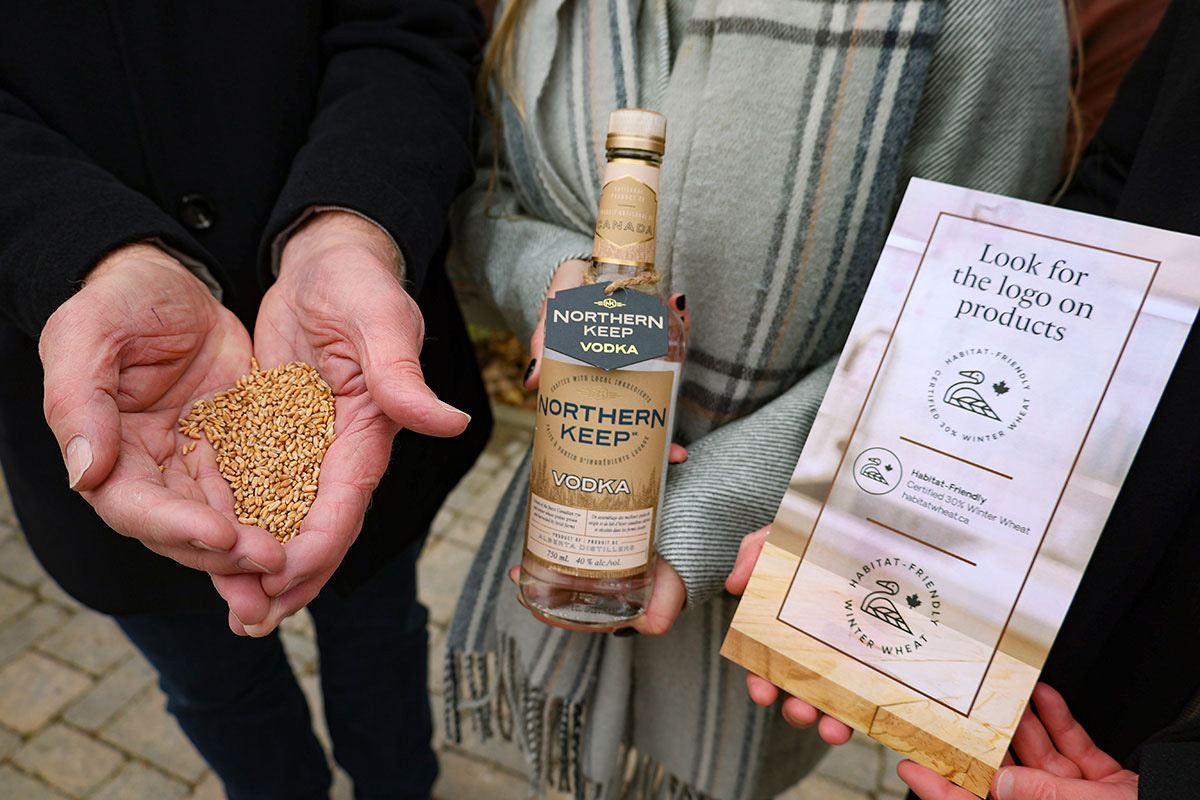 Three sets of hands holding out three separate items. The items are a handful of grain, a bottle of Northern Keep Vodka, and a plaque that says "Look for the logo on products" and continues to describe Habitat-Friendly Winter Wheat.