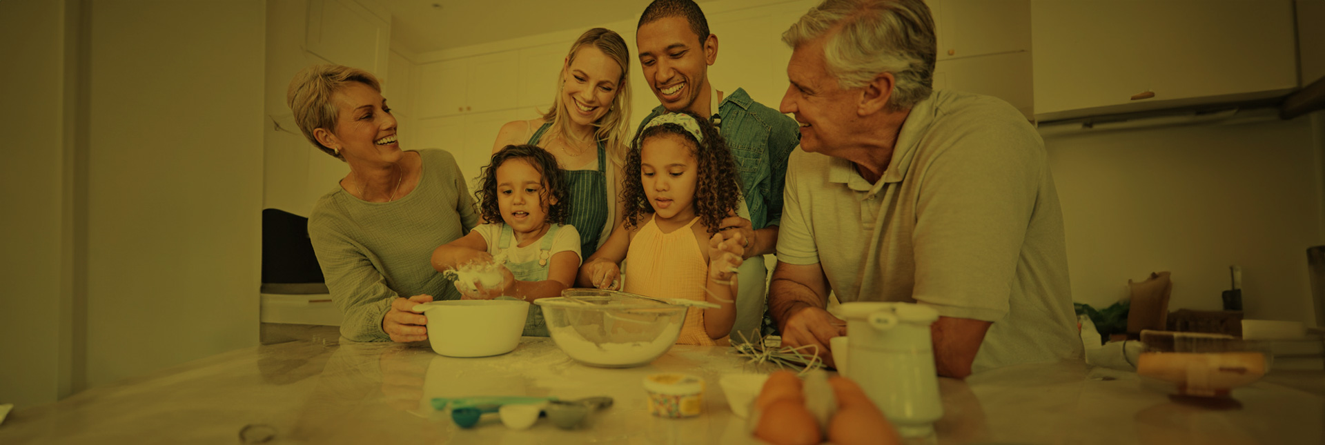 A multi-generational family groups together at their kitchen island. The kids have their hands in bowls of flour, and the parents and grandparents are assisting with the baking and are having a great time.