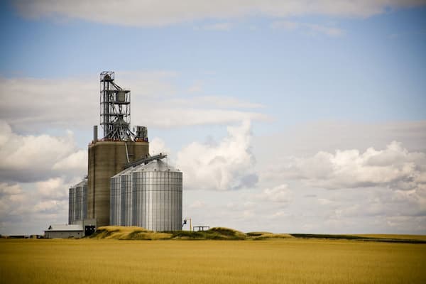 A grain elevator and large farming equipment and buildings stand on golden wheat fields, framed by the large blue prairie sky.