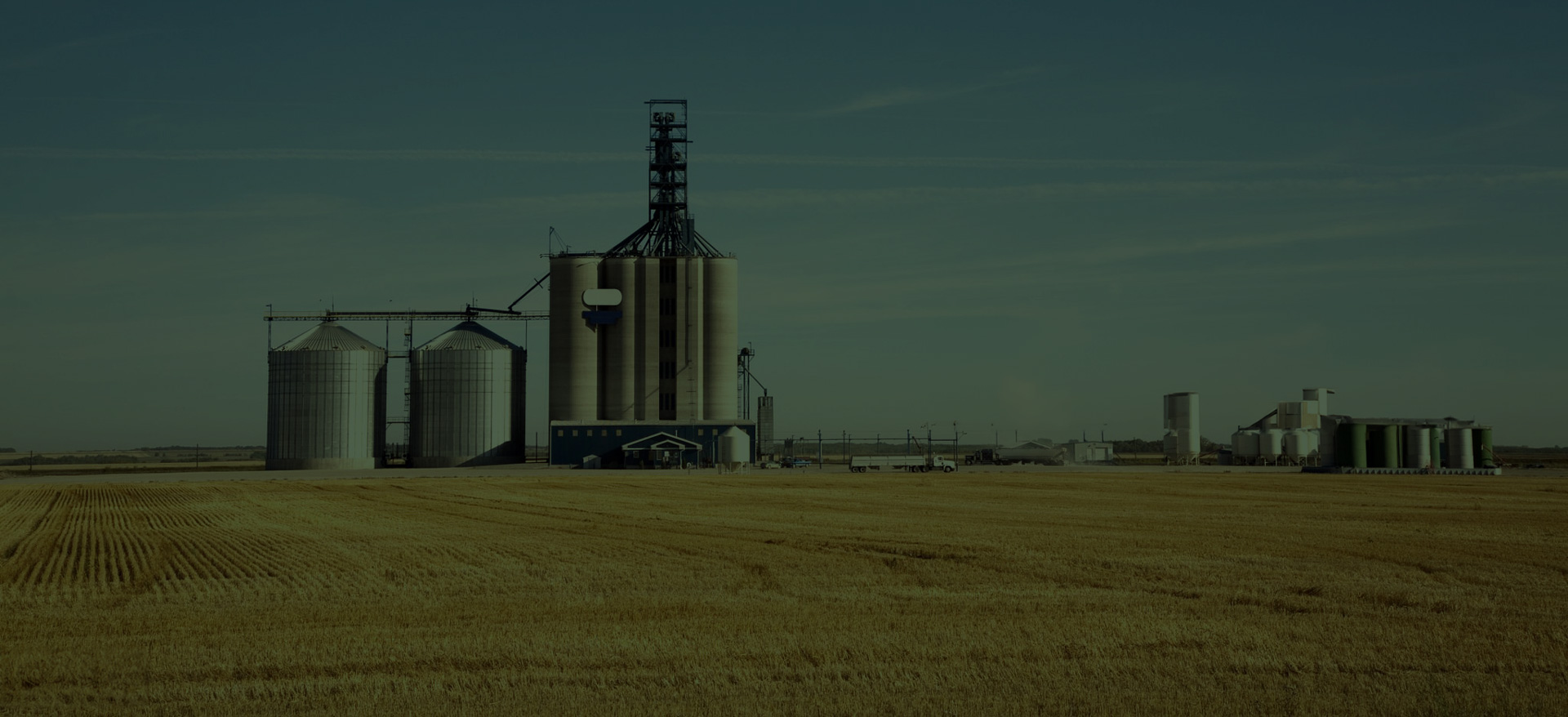 A grain elevator and large farming equipment and buildings stand on golden wheat fields, framed by the large blue prairie sky.