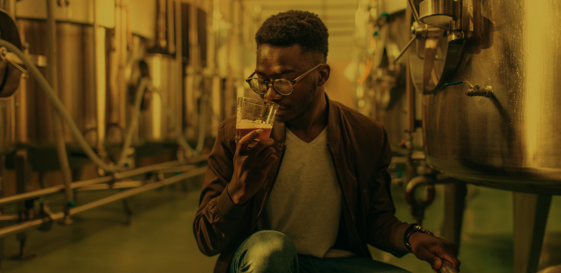 A man is kneeling next to a keg in a professional brewery. He is smelling a glass of beer.